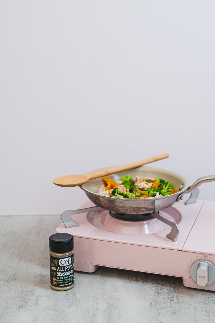 Pink portable stove with a wok containing vegetables and a wooden spoon, next to a container of All Pups Seasoning on a light surface.