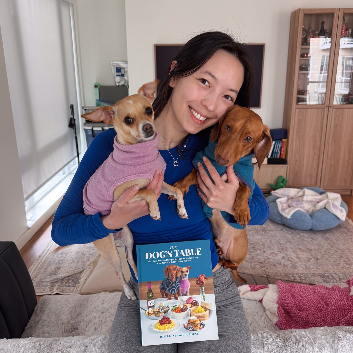 Joelle Jay holding famous dogs Cedric and Edith, and their cookbook titled 'The Dog's Table', in a home setting.