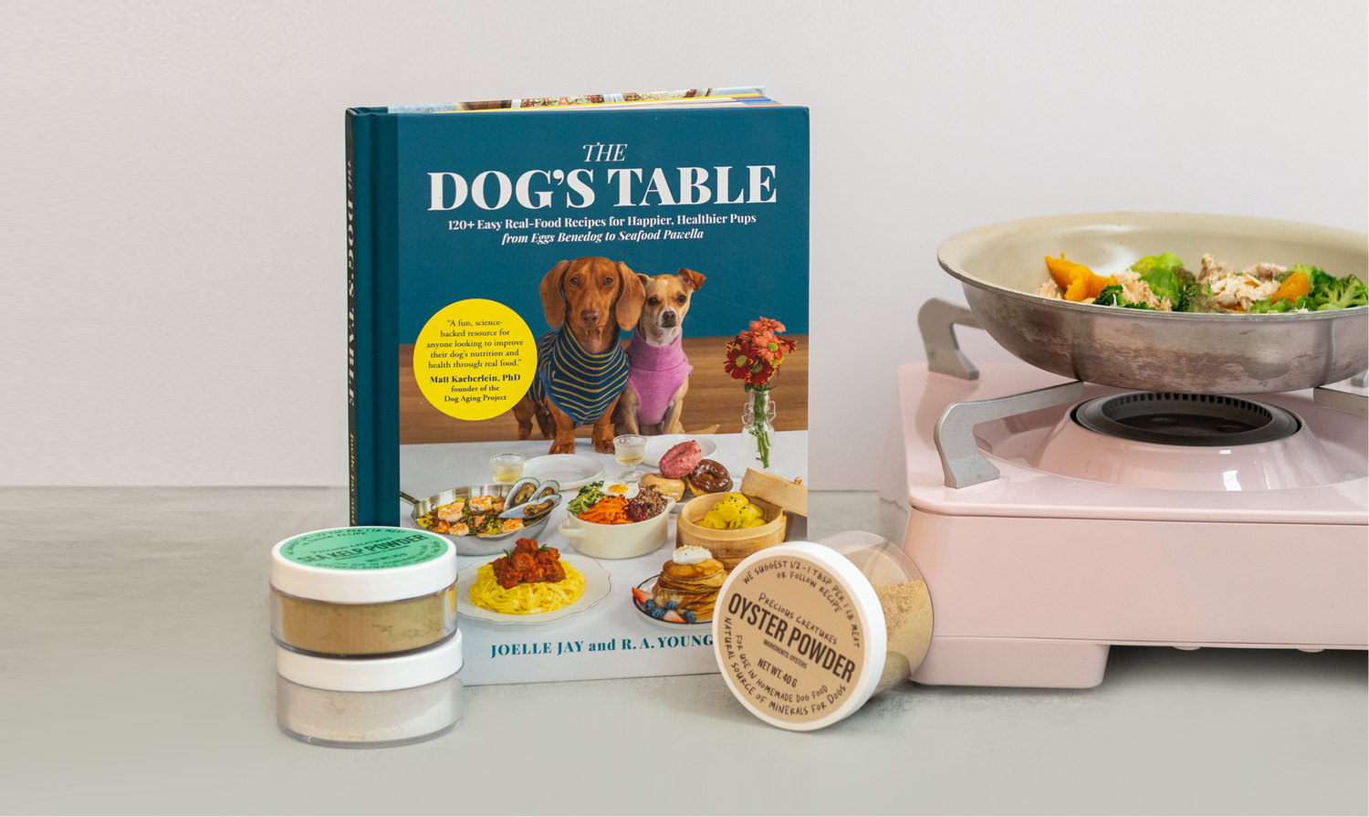 Book titled 'The Dog's Table' with Precious Creatures natural nutrient powders for homemade dog food and a pan of real dog food cooking on a stove.