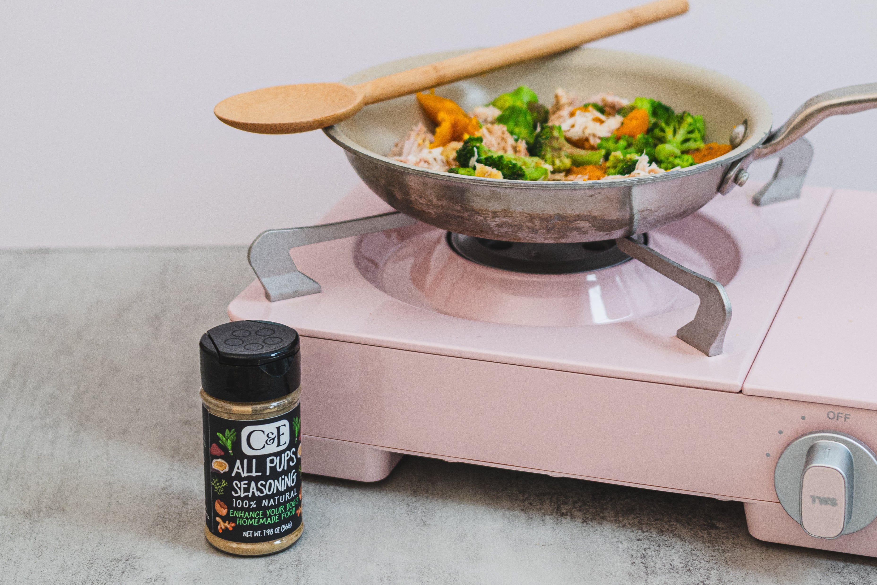 Pink portable stove with a wok containing vegetables and a wooden spoon, next to a container of All Pups Seasoning on a light surface.
