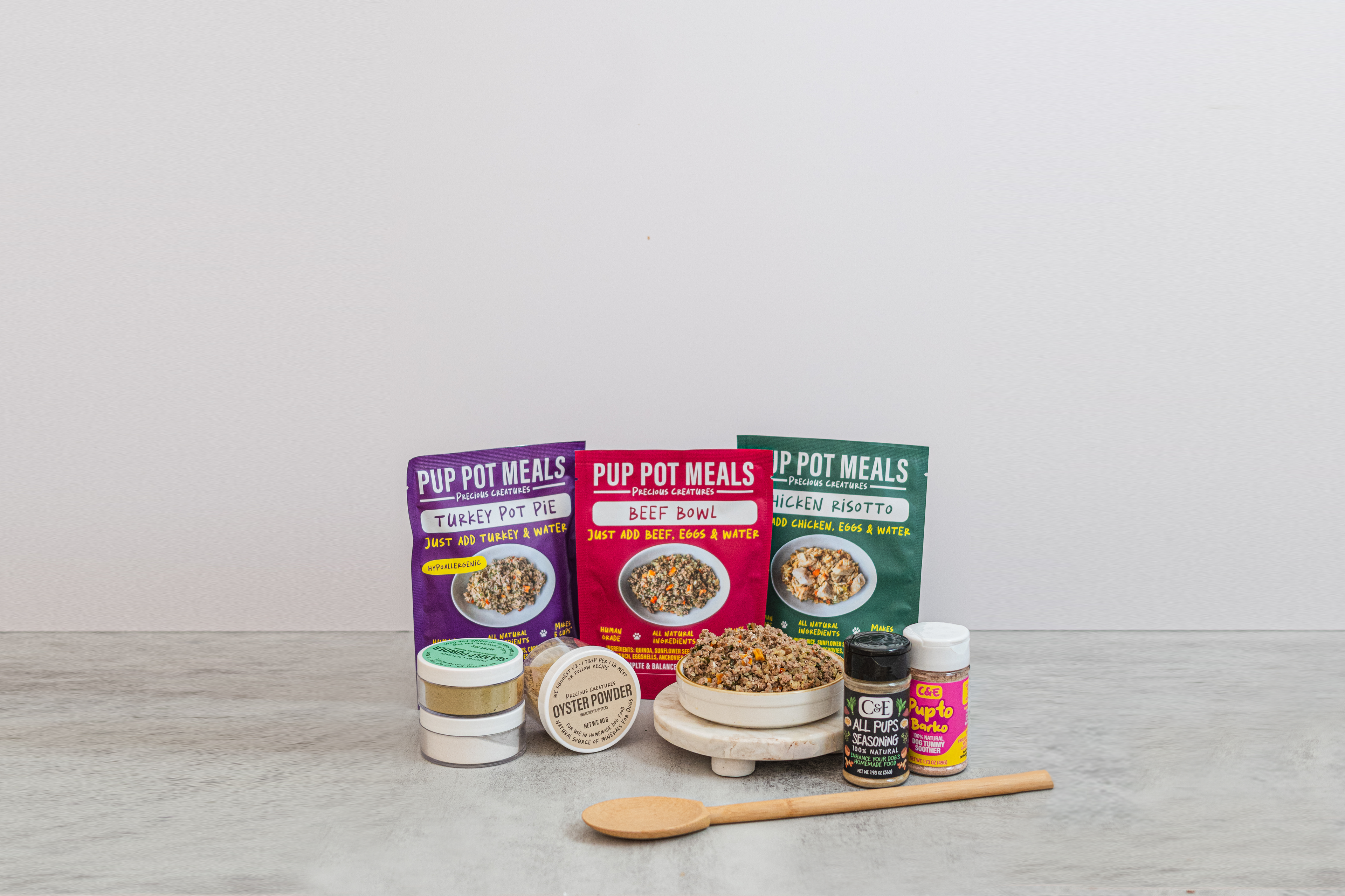 Assorted Precious Creatures Co homemade dog food supplement products including containers, bowls, and a spoon on a light gray surface with a white background.